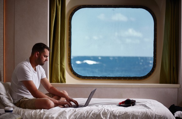 A young man on his laptop on his bed in a cruise ship cabin. 