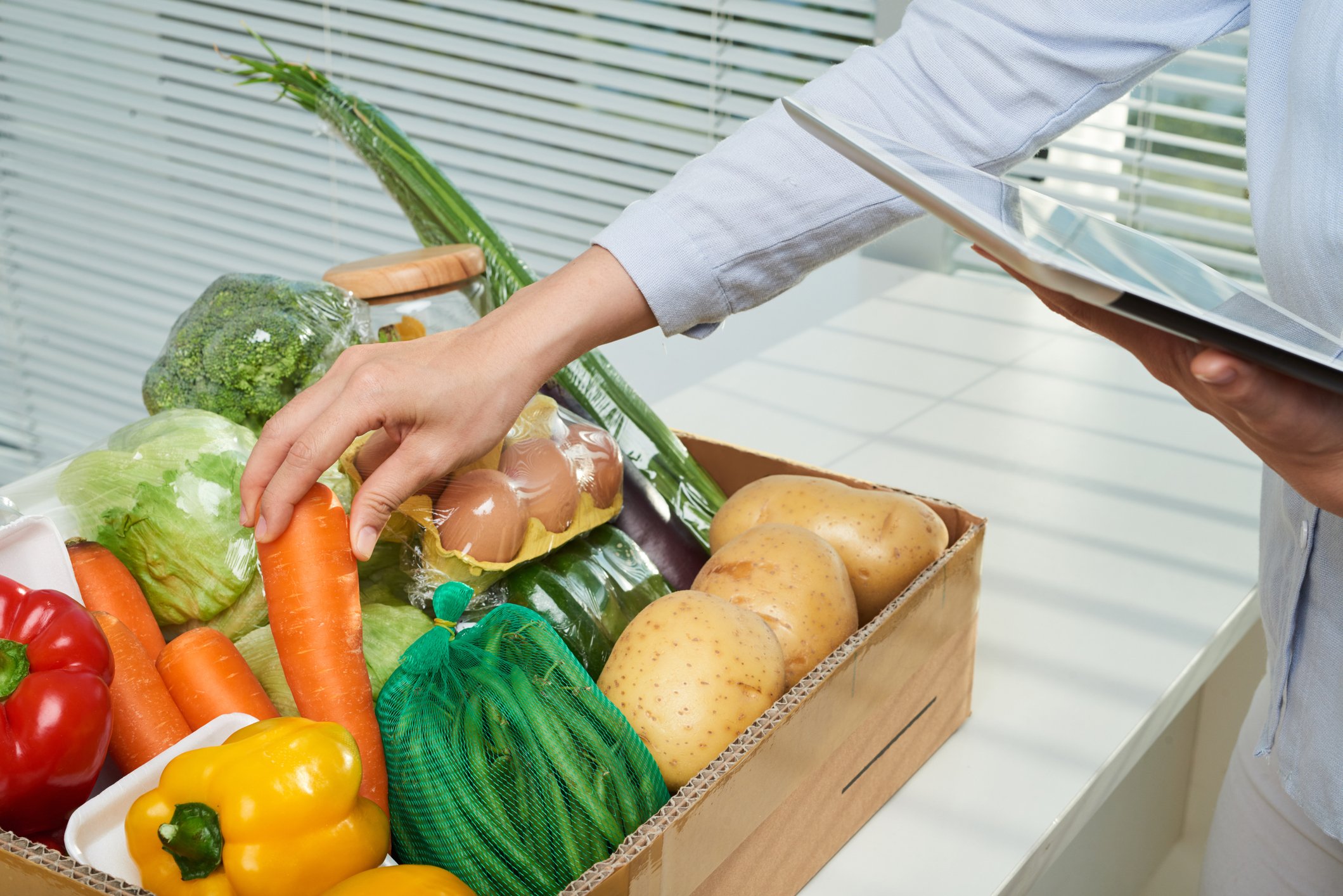 A woman grabbing a carrot from a box of vegetables as she checks her tablet.