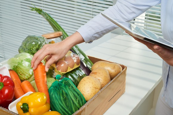 A woman grabbing a carrot from a box of vegetables as she checks her tablet.
