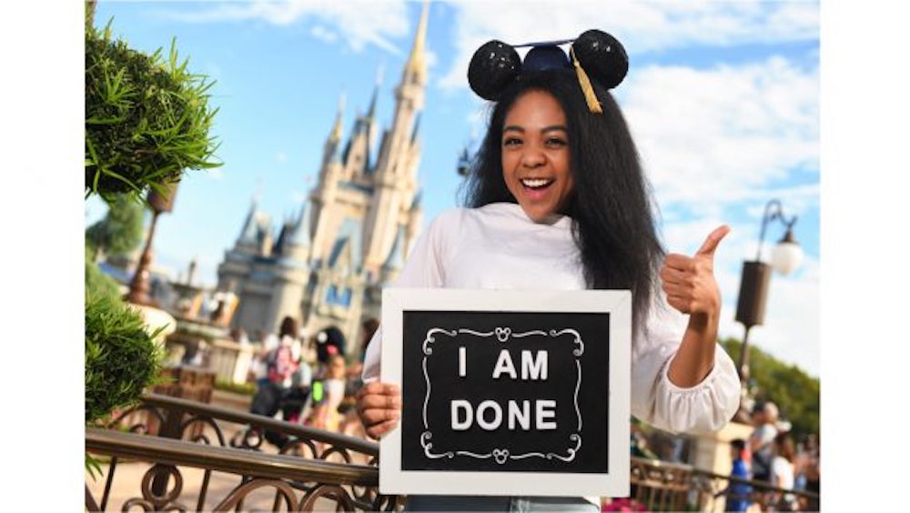 A high school graduate with an "I am done" sign as she wears mouse ears in front of the Magic Kingdom's Cinderella Castle.