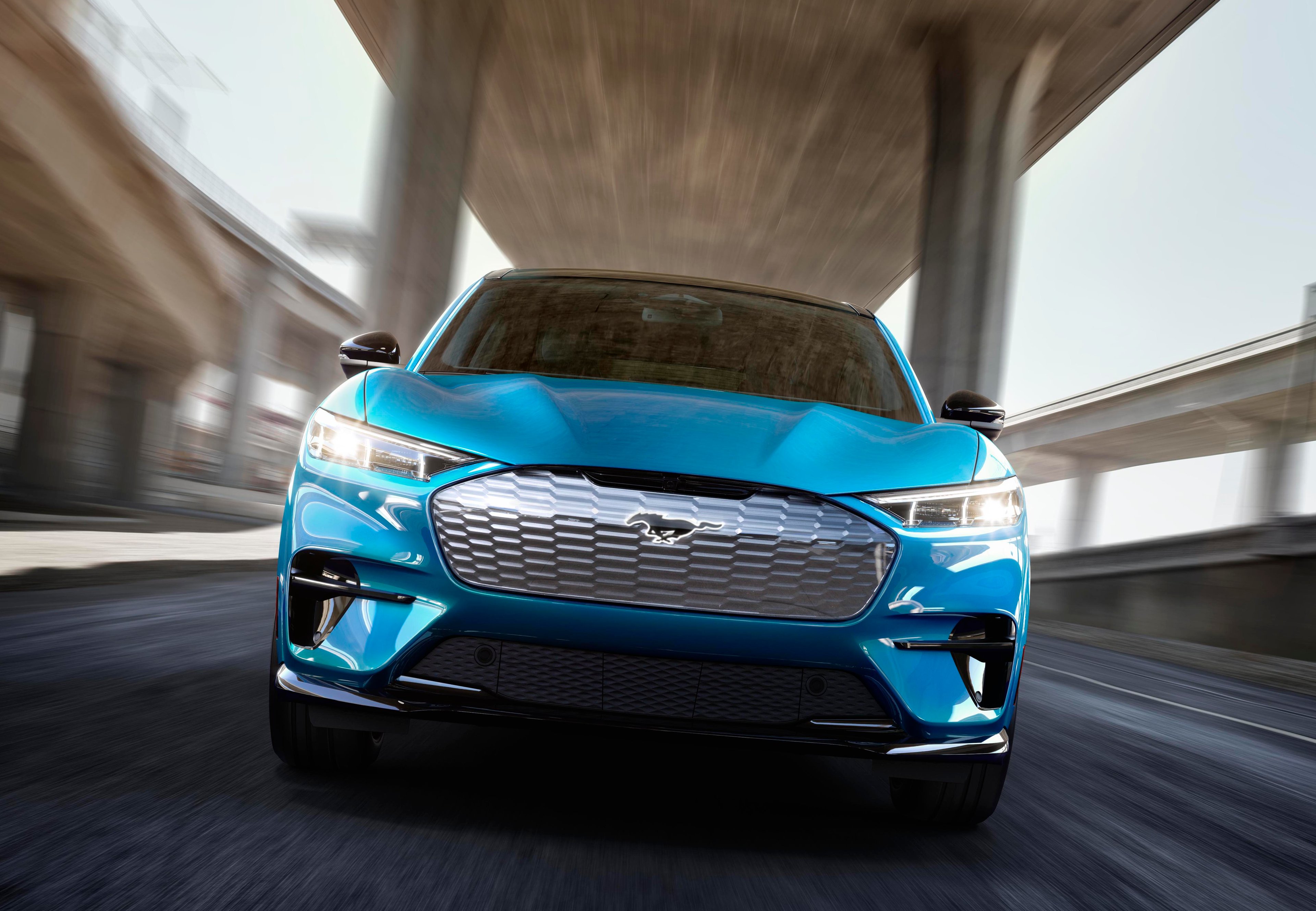 A blue Ford Mustang Mach-E, an electric high-performance SUV, shown from the front at speed on a city road.