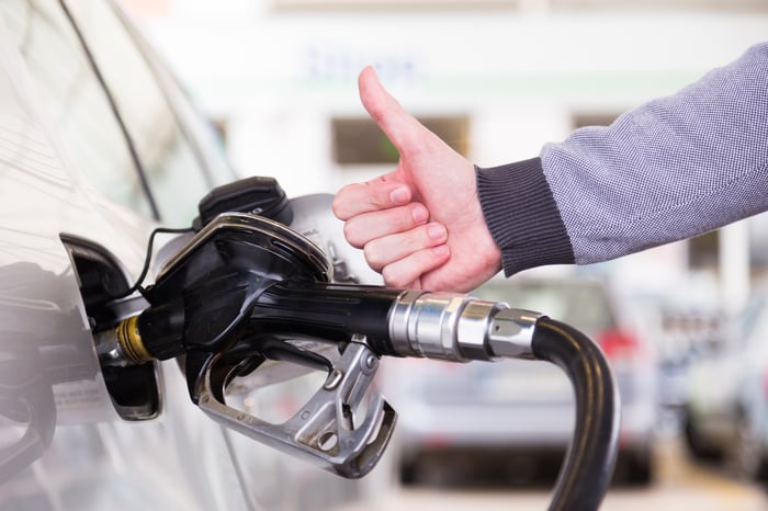 A gas pump refills a car's gas tank while a man's hand gives a thumbs-up.