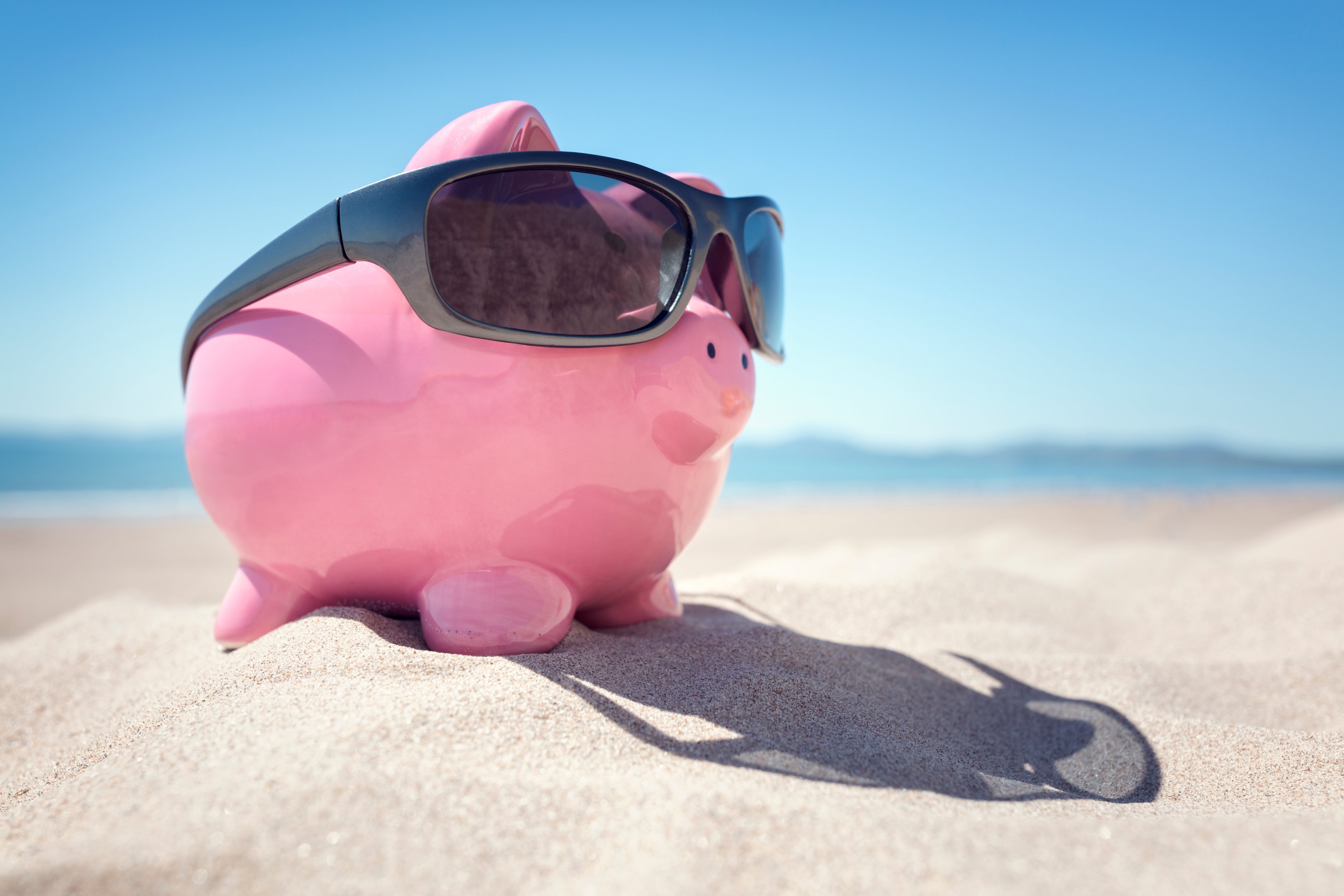 A pink piggybank with black sunglasses sits on a beach.