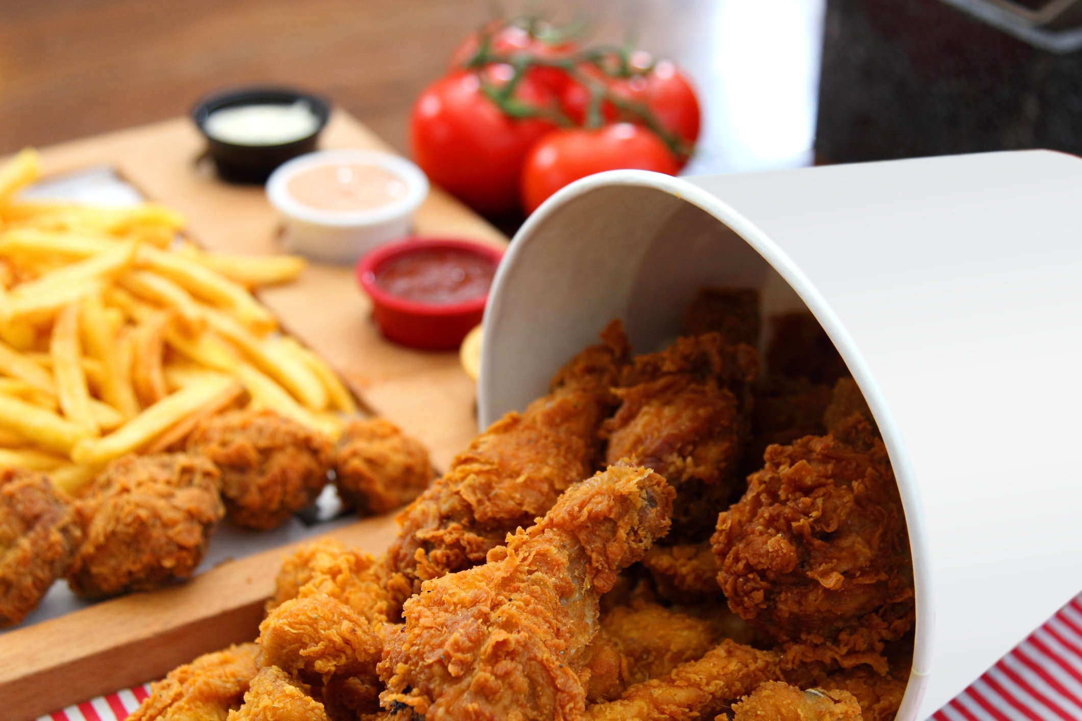 A bucket of fried chicken with fries and condiments in the background.