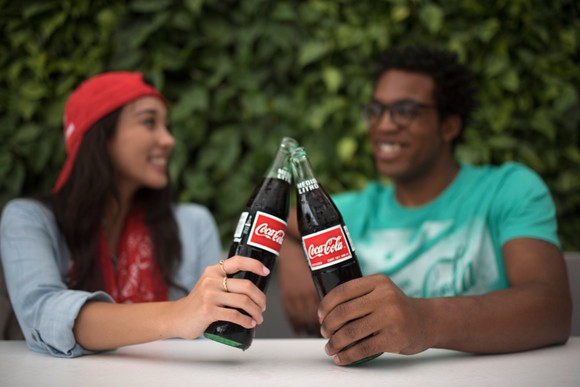 Two friends clanking their Coca-Cola bottles together while seated and chatting outside. 
