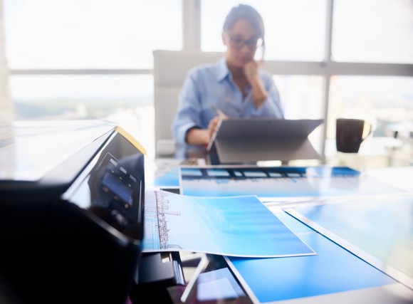 A woman working on a tablet computer with a printer sitting on a table.