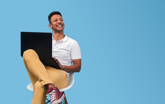 A happy-looking man sitting on a chair and looking up from his laptop.