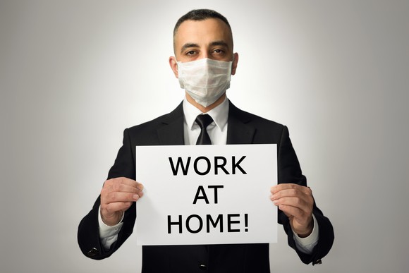 A man in a suit wearing a protective mask holding a sign that says "work at home!"