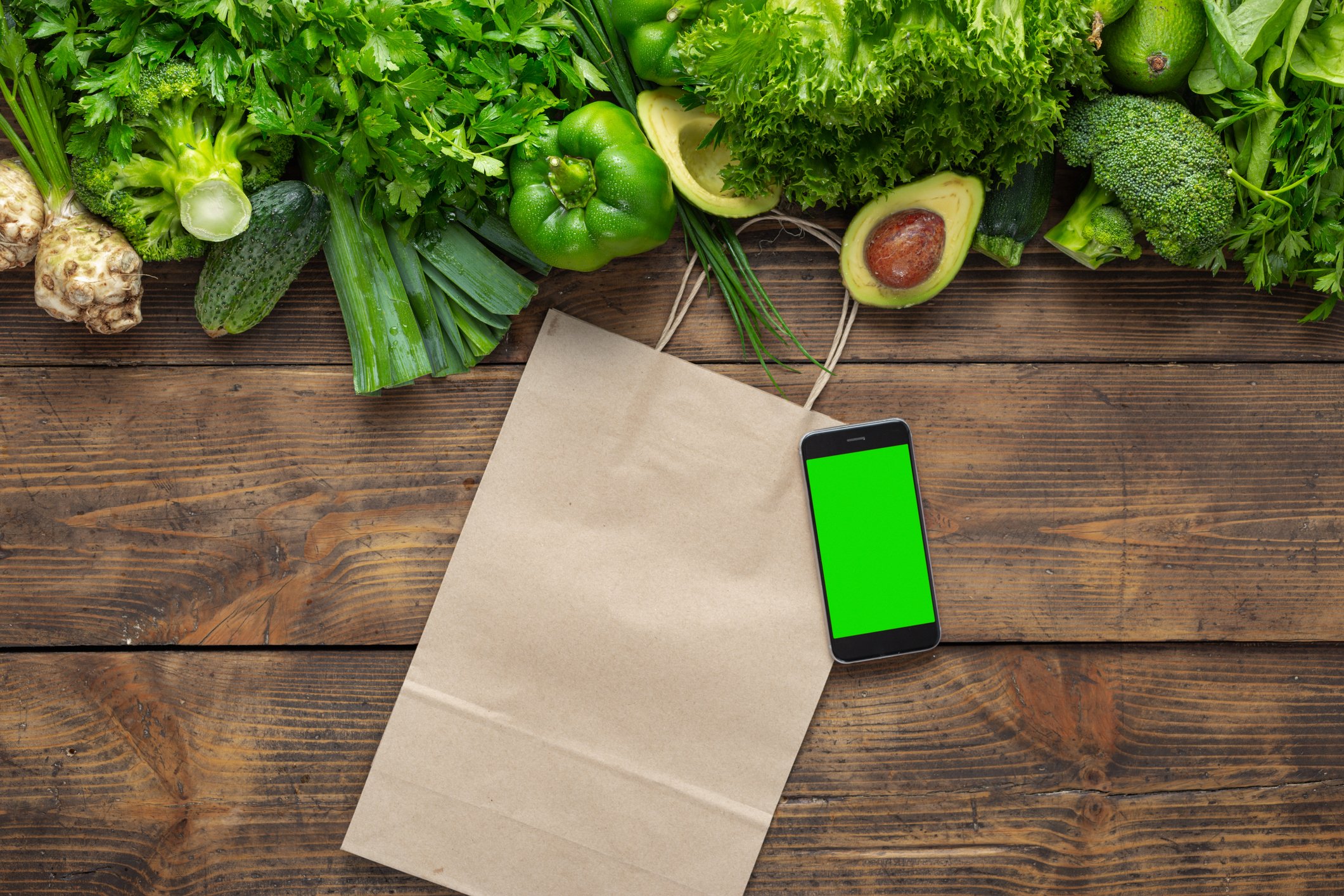 Green leafy vegetables are shown on a wooden surface with a paper shopping bag and a cellphone.
