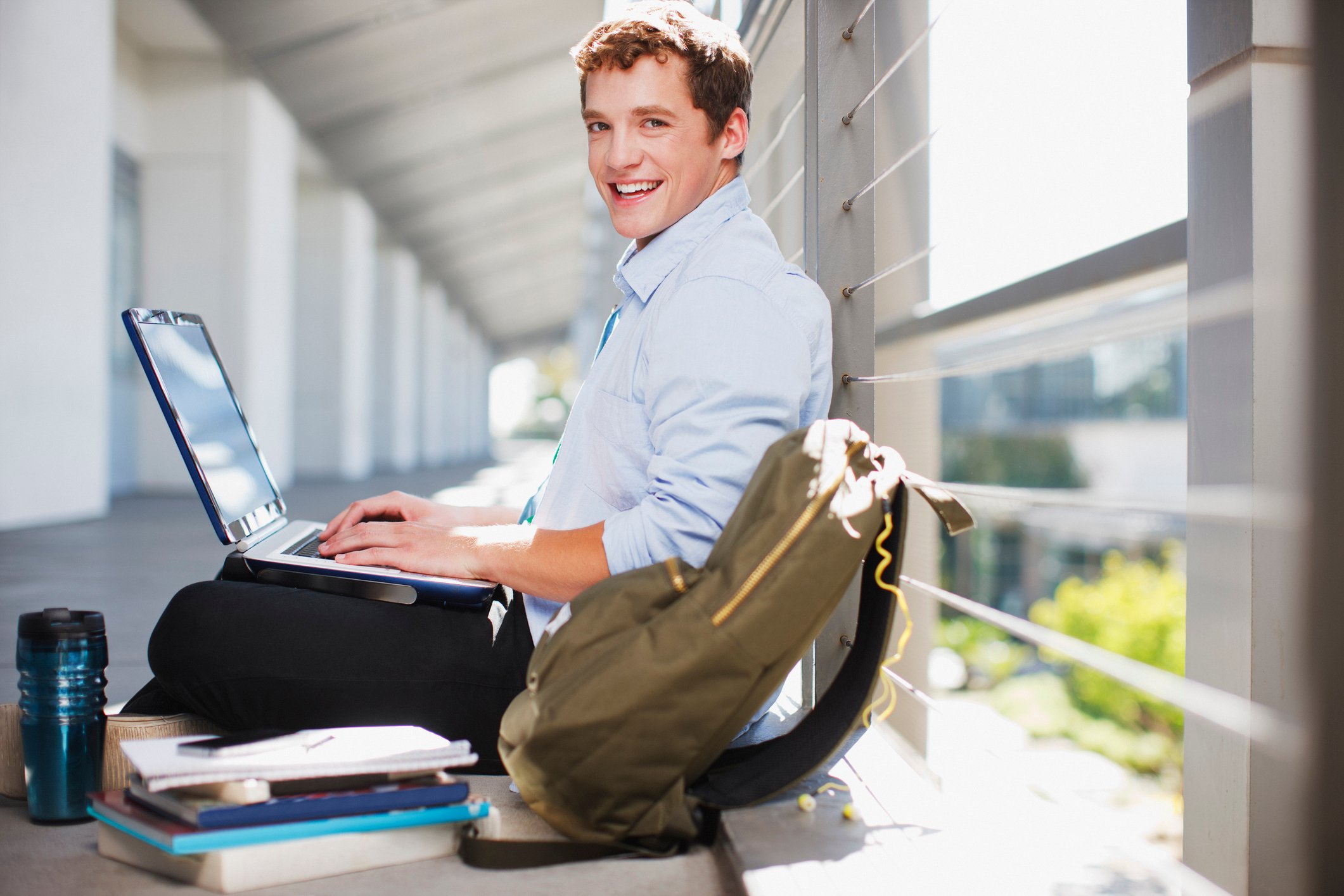 College student sitting on a floor with his backpack beside him and working on laptop.
