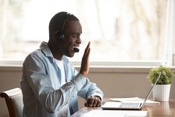 A teacher greeting his students with a wave from his home in front of his laptop with headphones and a microphone.