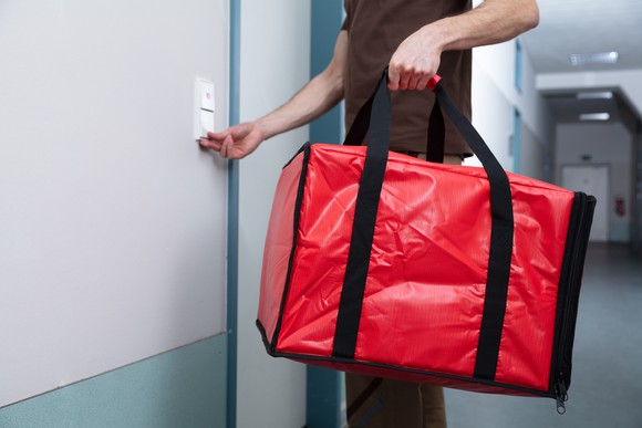 A delivery person rings a door bell while carrying a red bag containing meals.