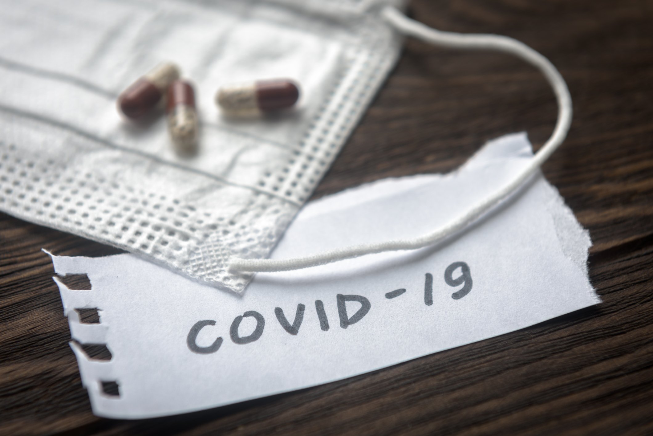 Pills and a mask are set on a wooden surface with 'COVID-19' written on a piece of paper.