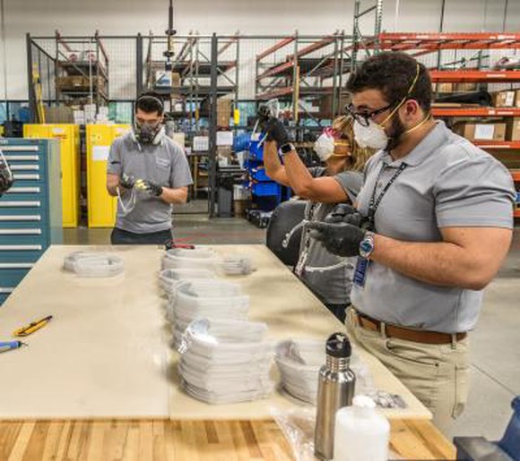 Three employees work at a table assembling face shields