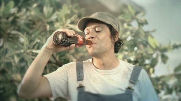 A man wearing overalls who's drinking a Coca-Cola. 