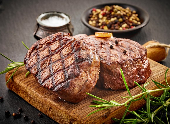 Two grilled steaks sit on a wooden serving board, flanked by condiments and sides.