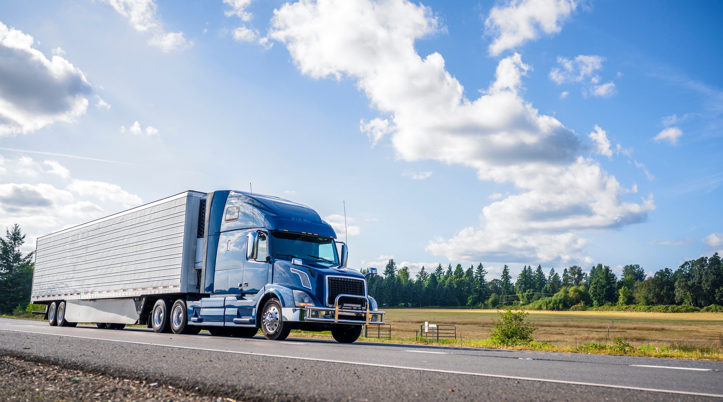 A refrigerated semi truck driving along a tree-lined highway.