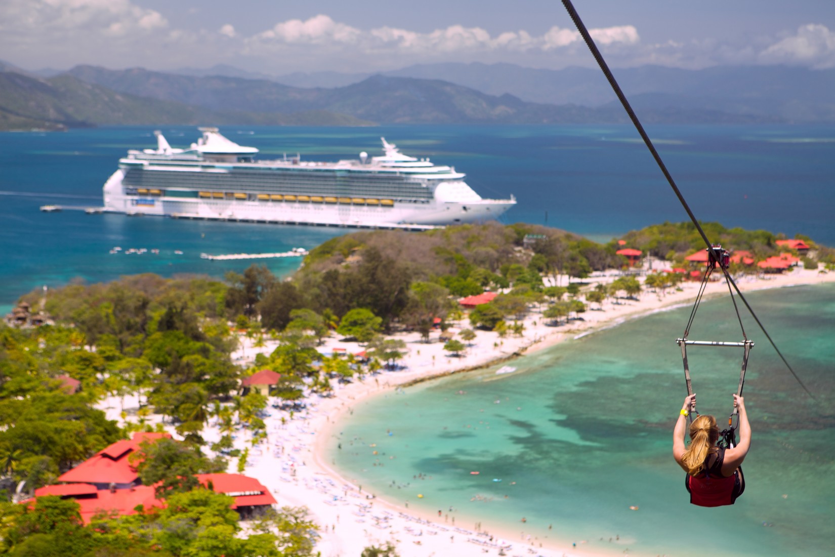 A woman ziplining along the beach in Labadee as a Royal Caribbean ship is docked in the background.