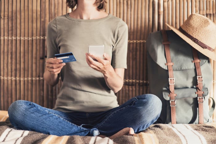 Woman sitting on a bed while using her credit card to make a purchase on her phone.