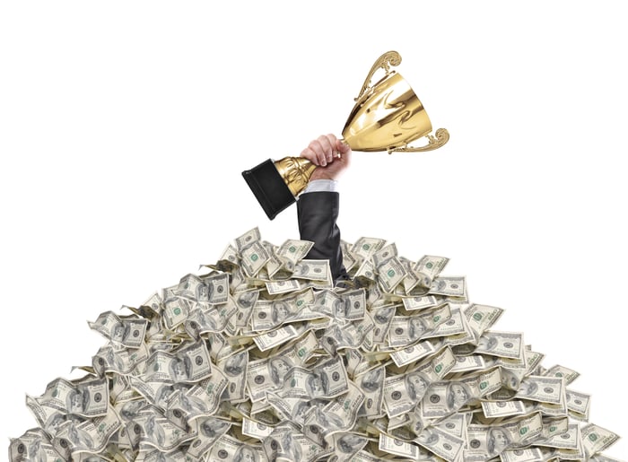 Hand holding a trophy sticking out of the top of a pile of $100 bills