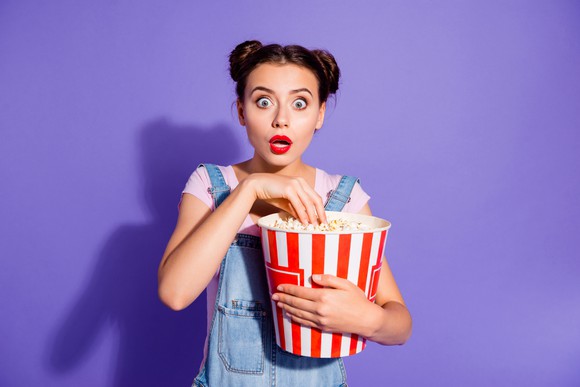 A young woman with a surprised expression on her face dips into a very large bucket of popcorn.