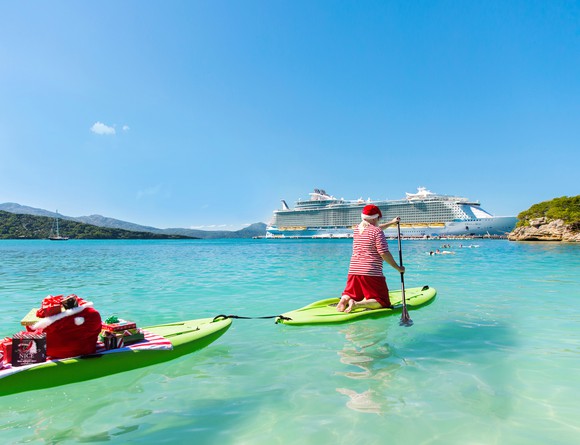 A paddle boarder dressed as Santa with presents in two paddles toward a Royal Caribbean ship.