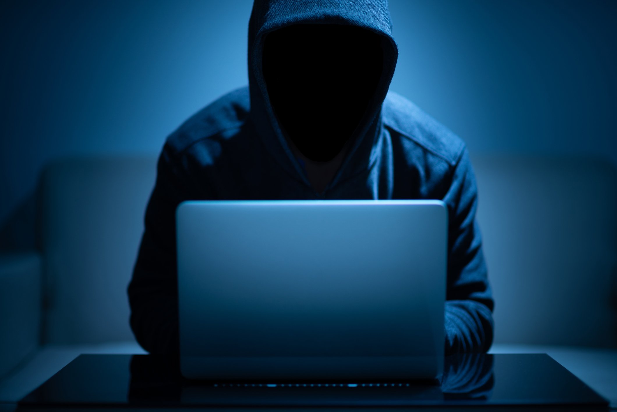 A shadowed man in a hoodie sits in front of a laptop.