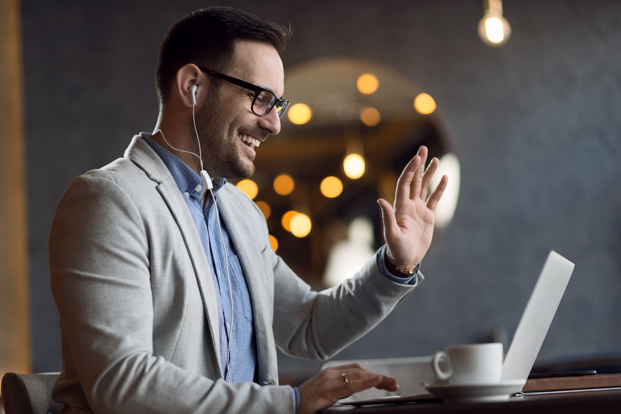 Man with a beard and glasses waves to his laptop as he participates in a video conference.