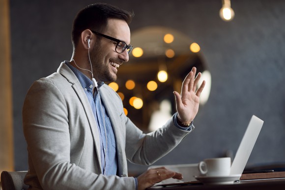 Man with a beard and glasses waves to his laptop as he participates in a video conference.