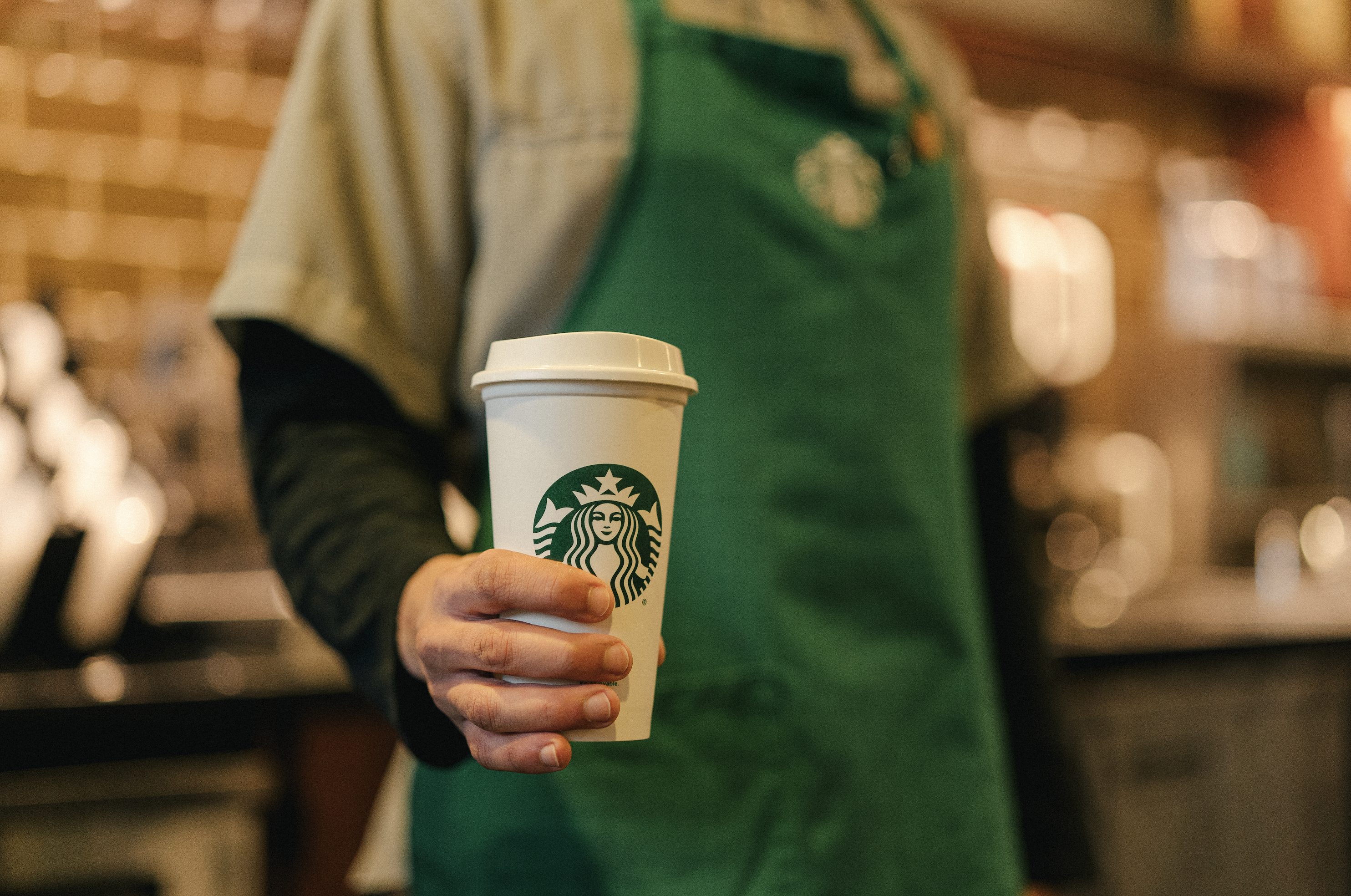A Starbucks employee, wearing a green apron, holds a cup with the Starbucks logo printed on the side.