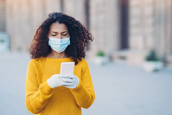 Woman looking at her smartphone while wearing a medical mask and gloves and standing outside. 