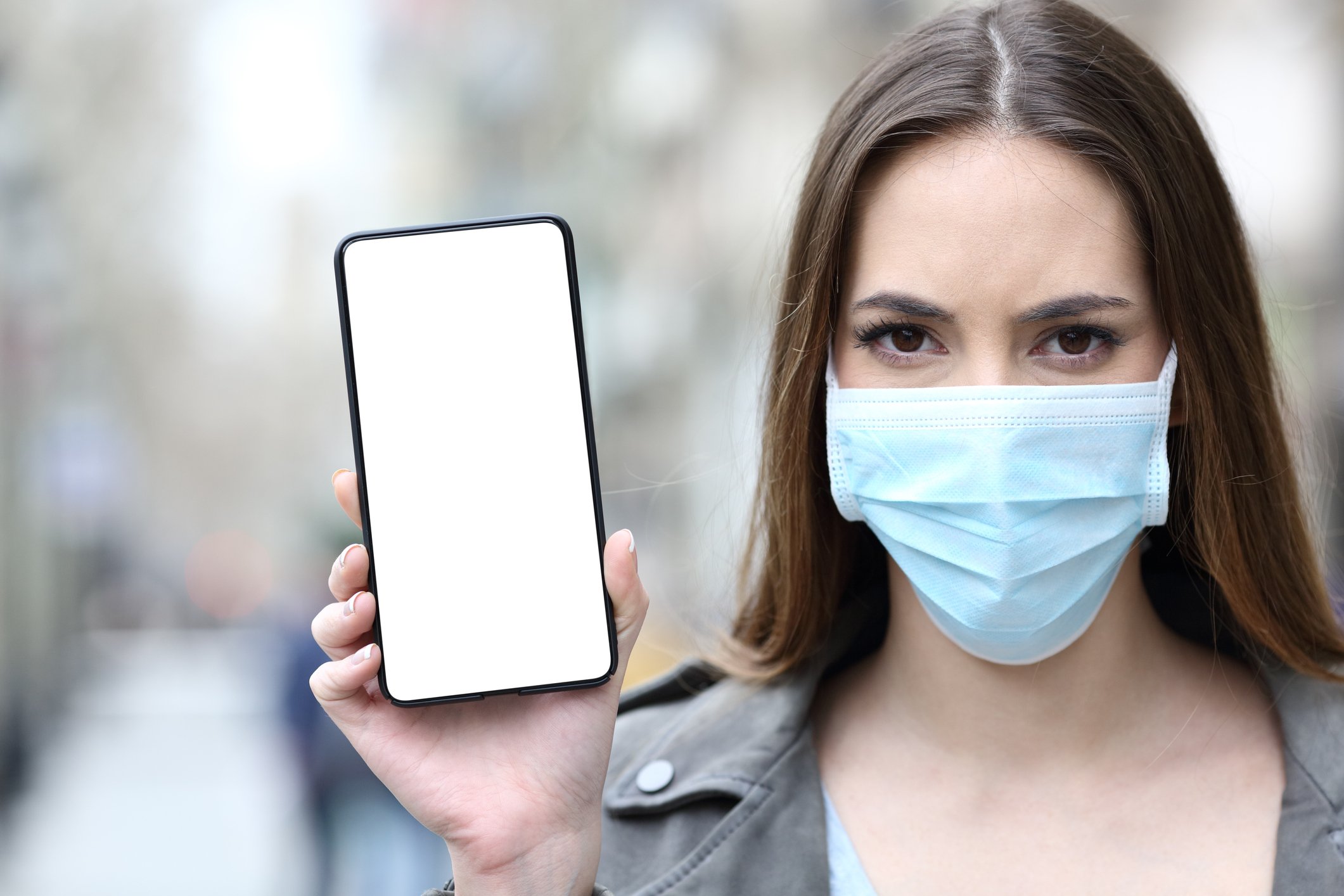 A young woman wears a face mask and holds up her smartphone while looking into the camera. 