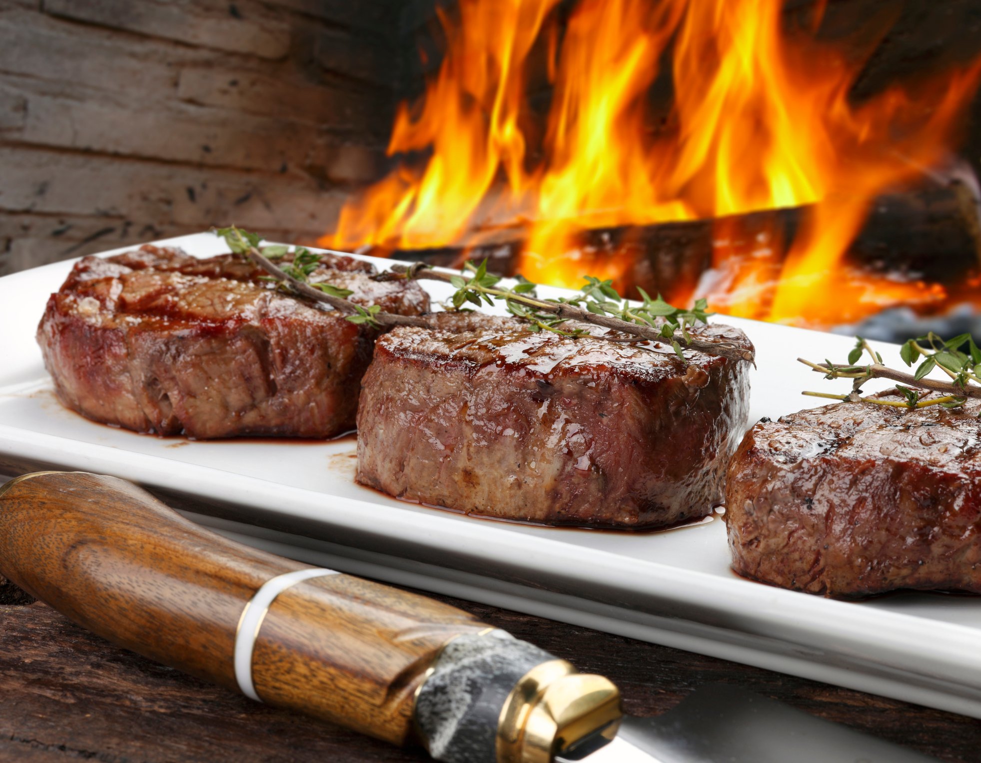 Three beef steaks on a platter beside a wood fire