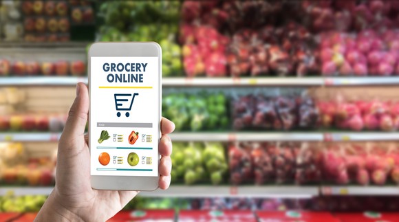 A hand holds a cell phone with "grocery online" written on the screen. Shelves of apples are seen in the background.