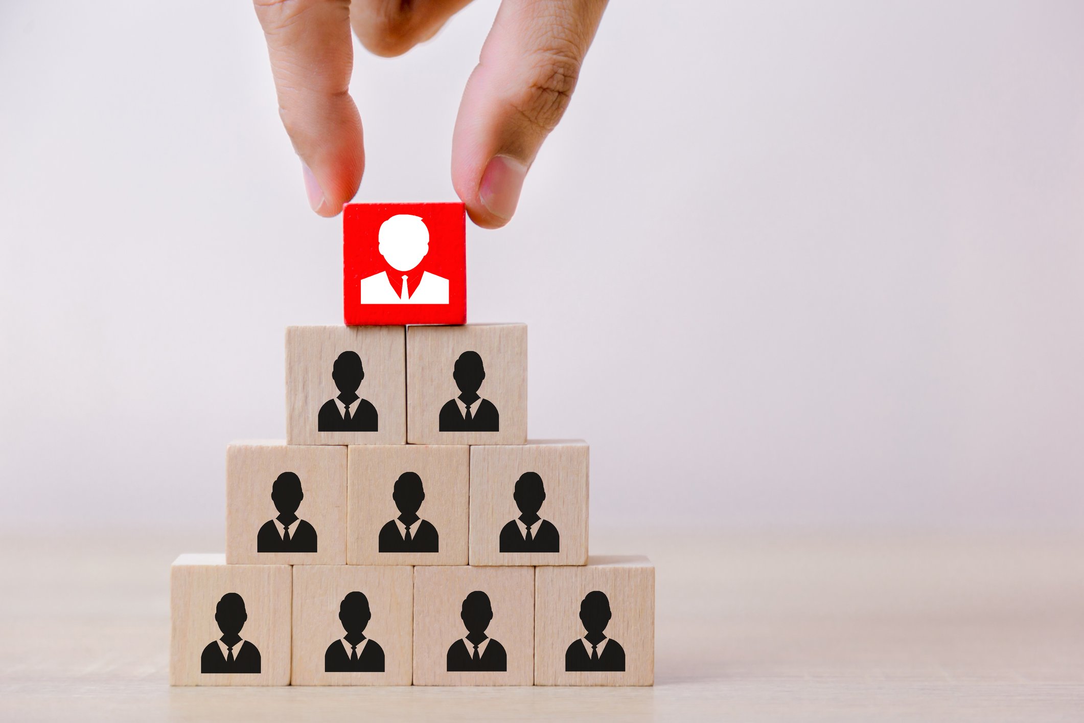 Hand placing red block on pyramid of blocks, each marked with the image of a person in a business suit, to represent a change in corporate hierarchy.