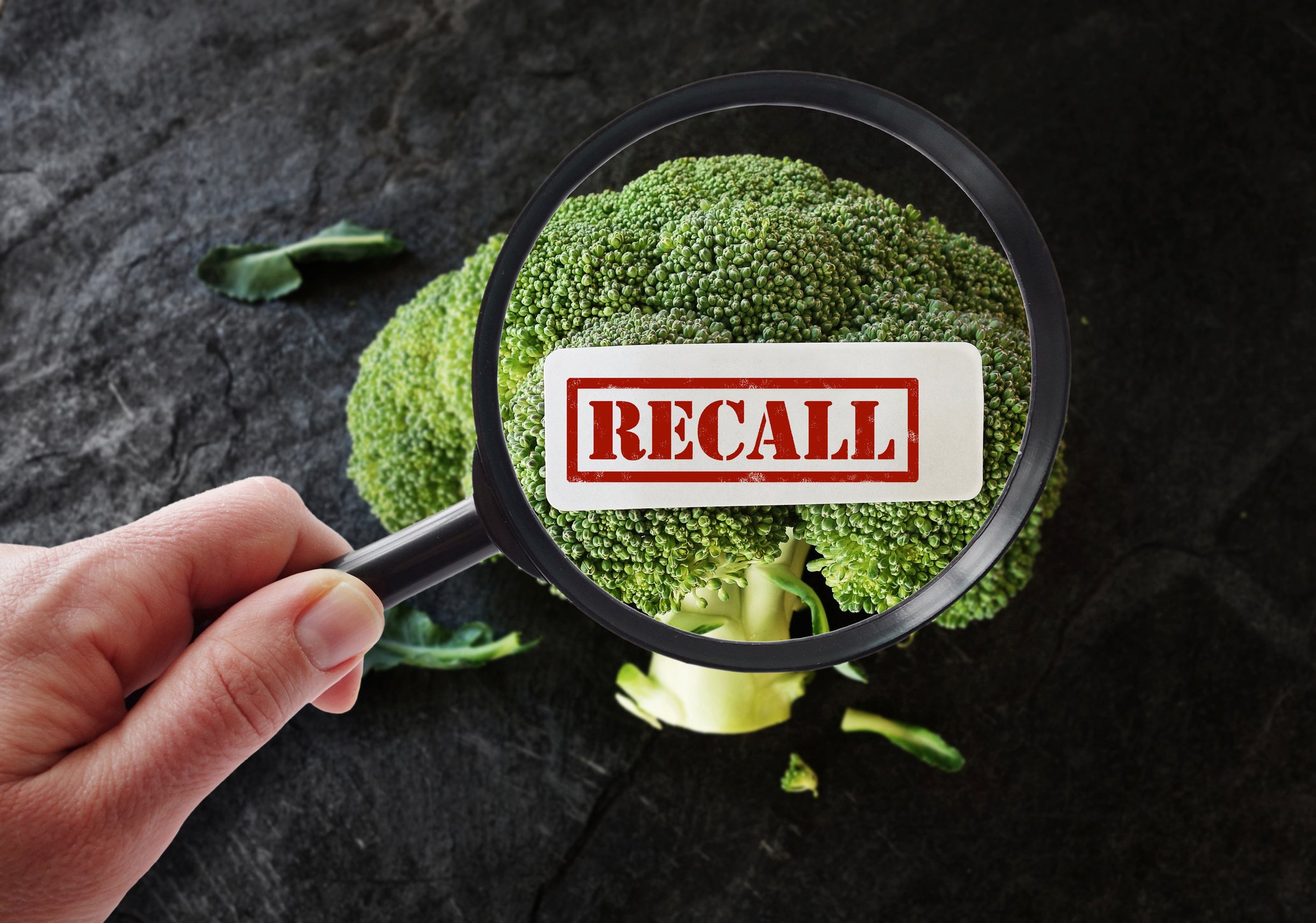 A hand holding a magnifying glass, looking at broccoli, with a Recall label superimposed.