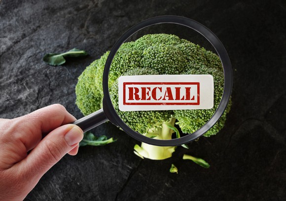 A hand holding a magnifying glass, looking at broccoli, with a Recall label superimposed.