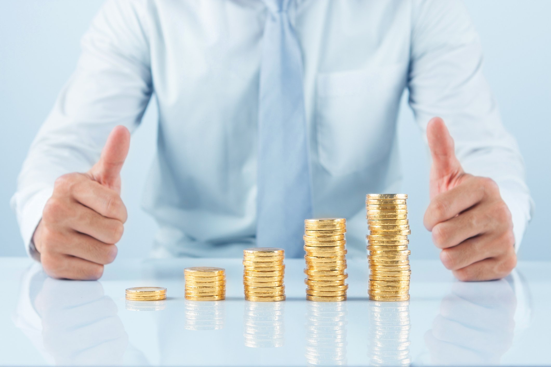 A person giving the thumbs-up sign while behind an ascending stack of coins.