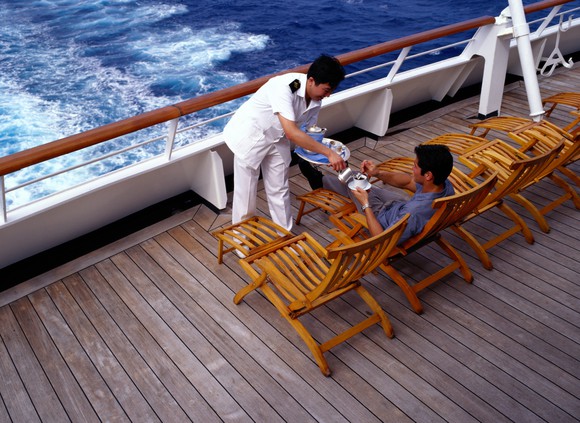 A waiter on a cruise ship serves coffee to a passenger lounging on the back. 