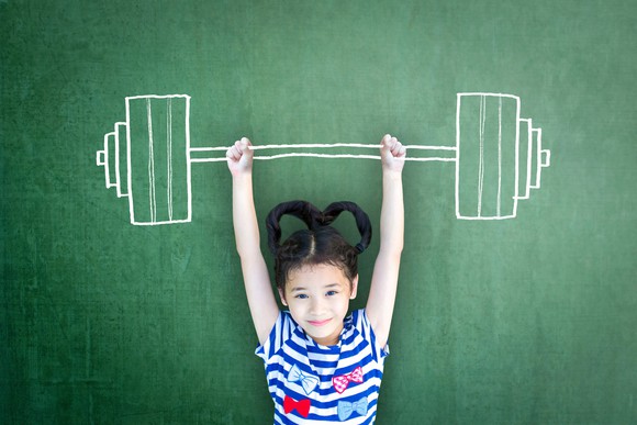 Small kid pretending to lift a large barbell that's drawn on a chalk board.