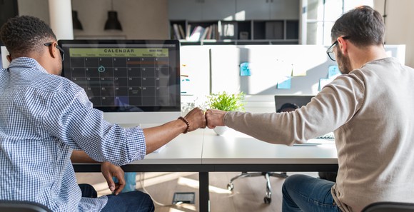 Two men touching fists looking at computers