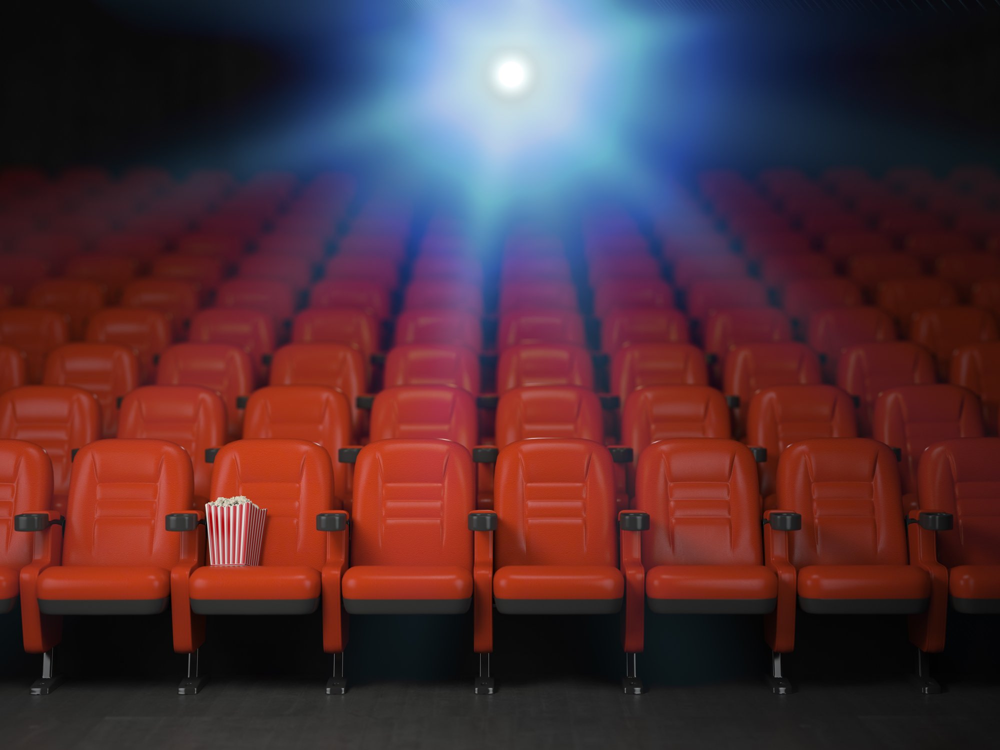 Empty theater seats, a box of popcorn in one of the seats, and a projector light in the background.