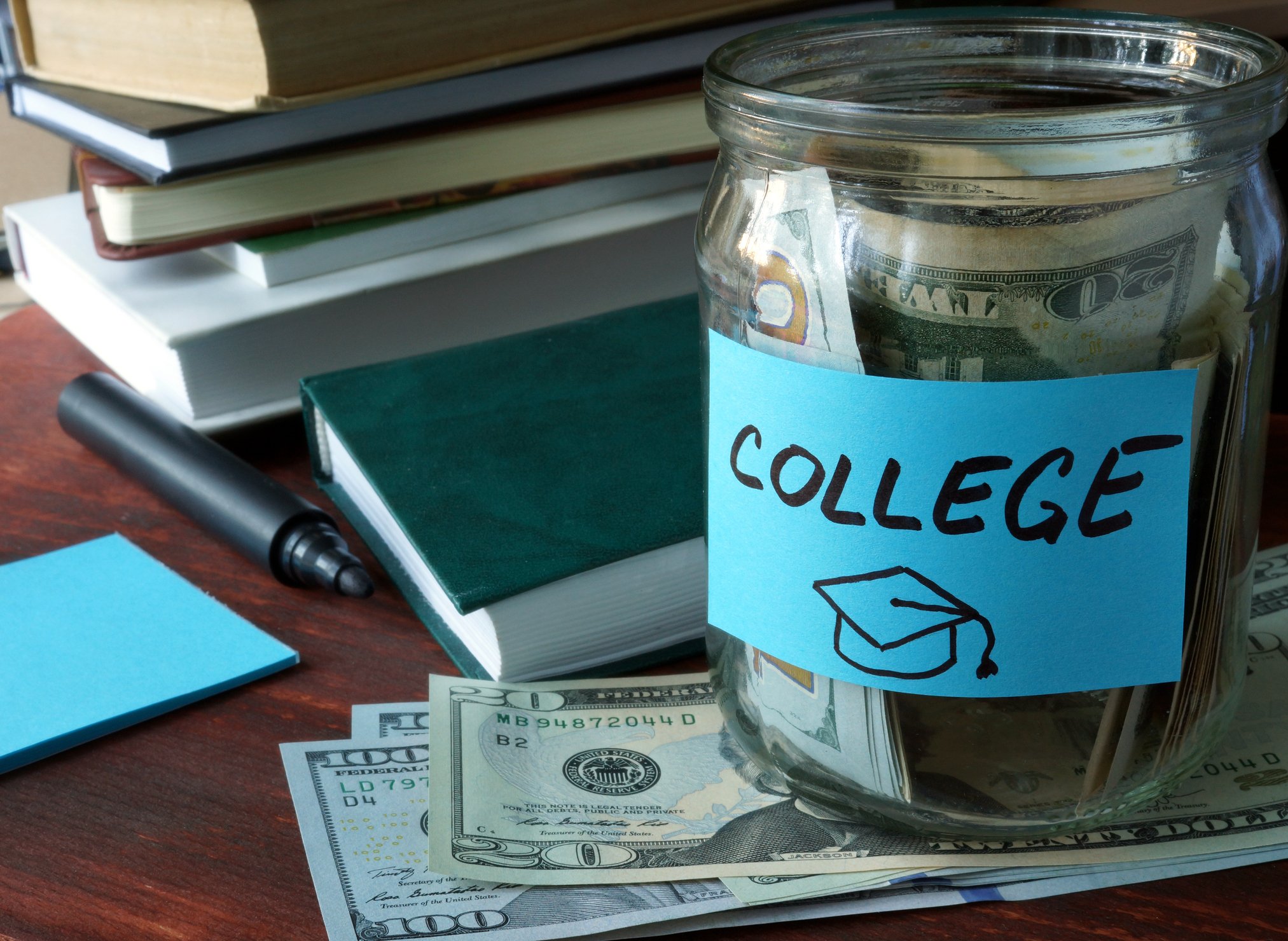 Jar filled with bills labeled college on table with bills under it, sitting next to books, a pen, and a notepad