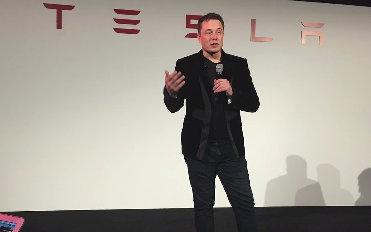 Person standing on a stage with a microphone in front of Tesla logo.