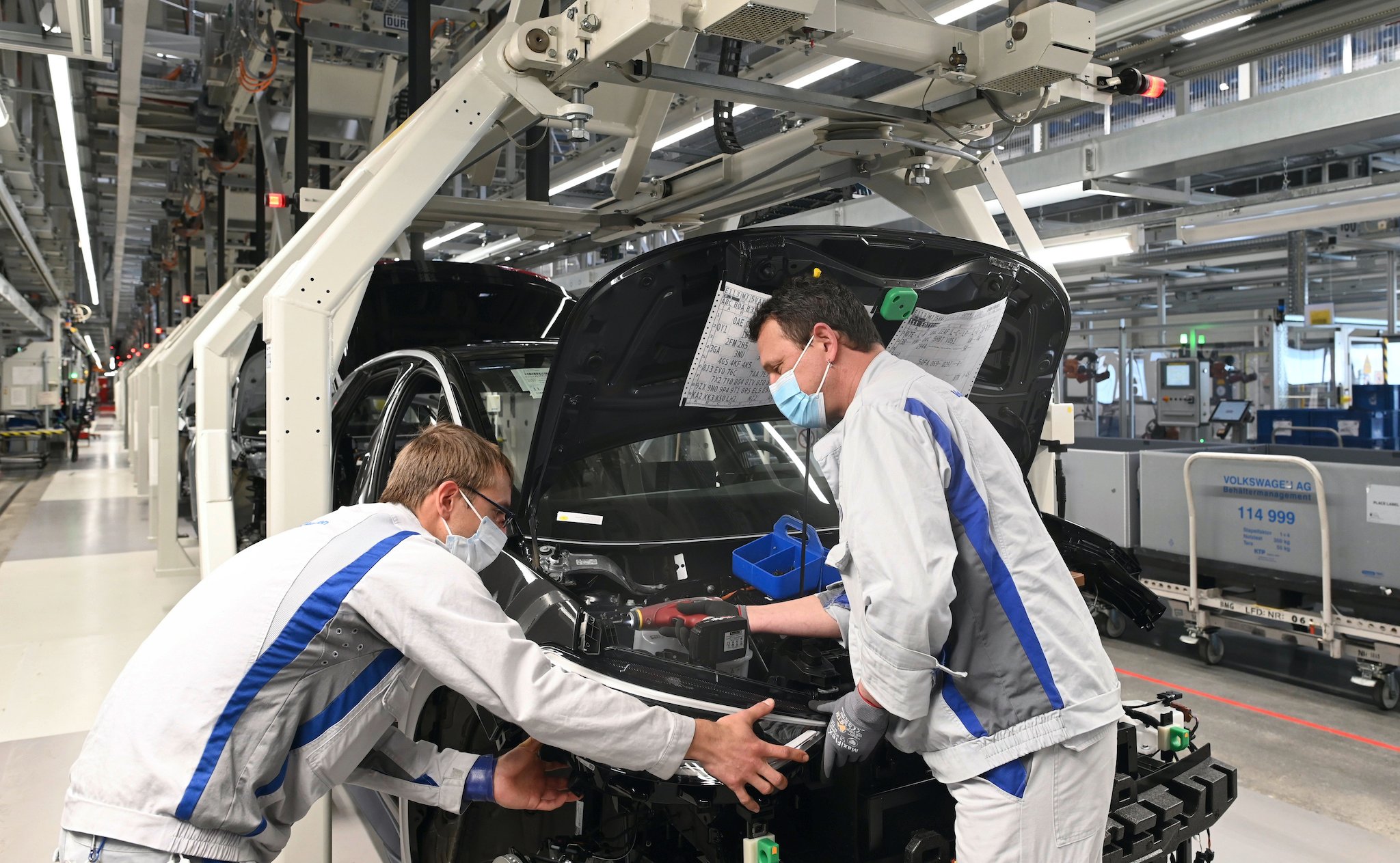 Two workers wearing masks tend to a partially-assembled VW ID.3 hatchback on an assembly line.