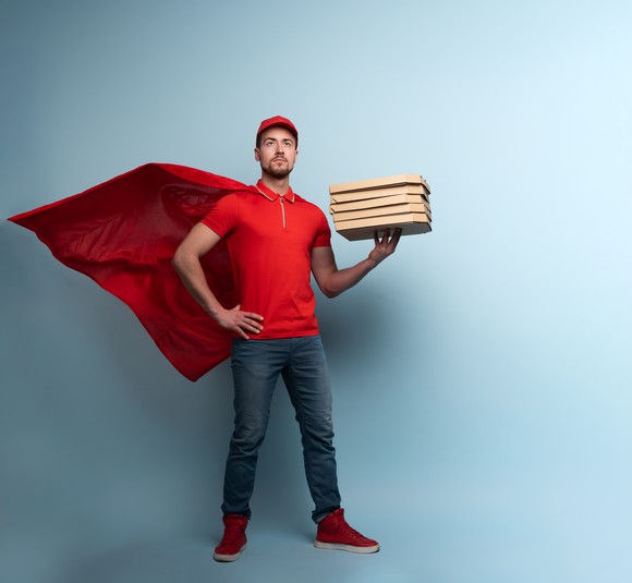 A man wearing a red cape and cap is holding 5 pizzas in his left hand.