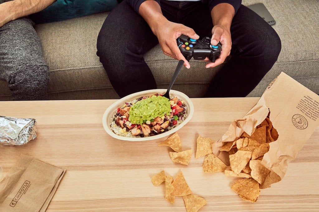 Man with a remote control in his hand sitting in front of a plate of food from Chipotle.