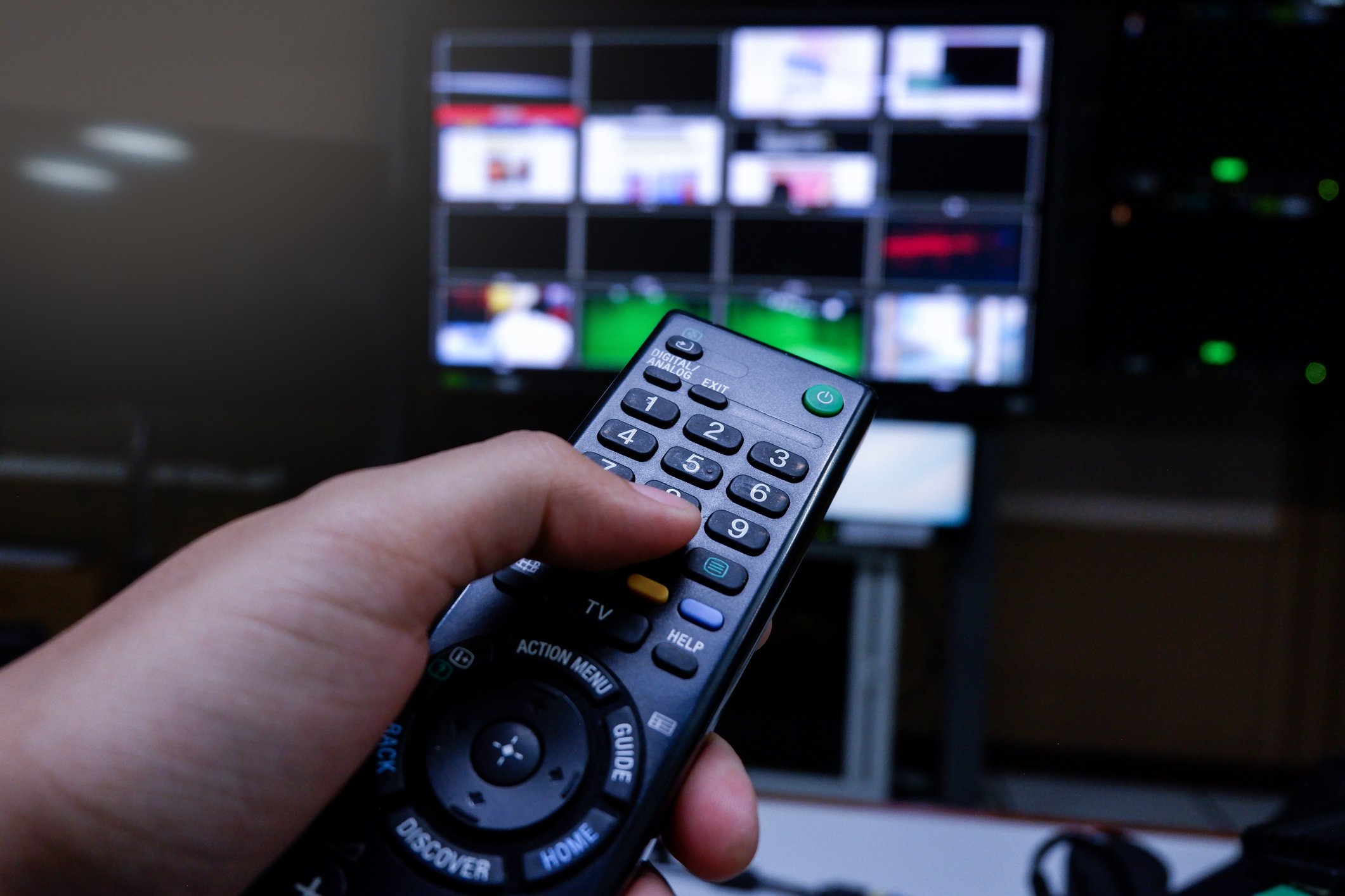 A hand holds a TV remote to change channels on a TV in the background of a dark room