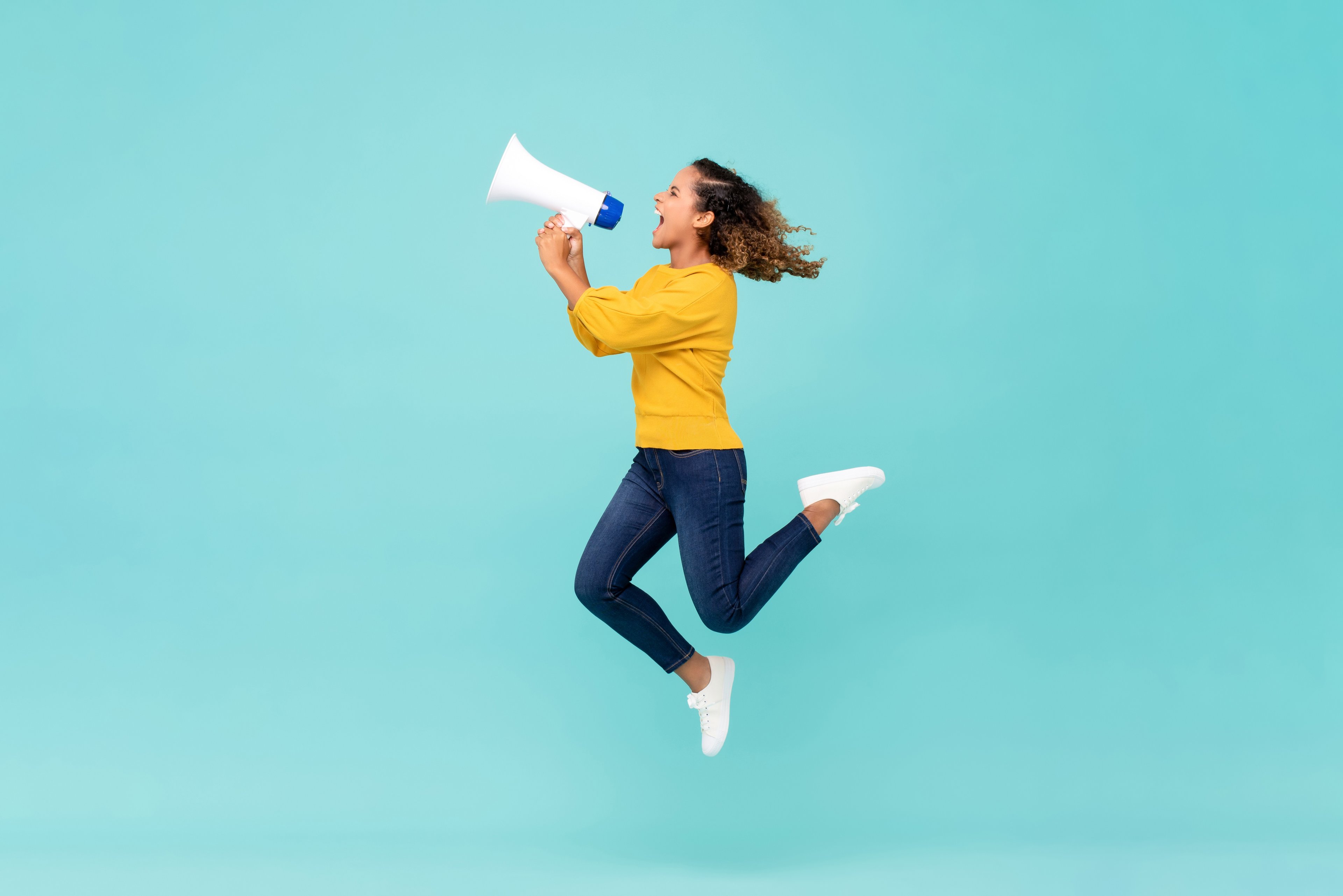 A women holding a bullhorn, wearing a yellow sweater, jumping to sell something. There's a light blue background. 