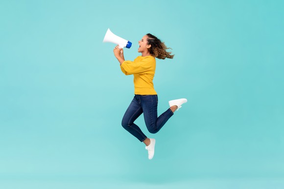 A women holding a bullhorn, wearing a yellow sweater, jumping to sell something. There's a light blue background. 